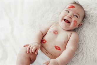 A laughing baby with red lipstick kisses on her face and body lies on a white cosy blanket