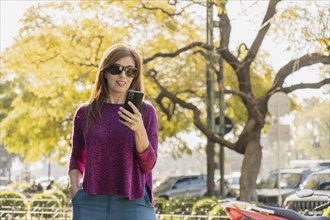 Stylish middle-aged woman smiles as she looks at her smartphone in a park at sunset. Wearing