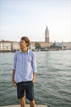 Young man in striped shirt and shorts on the banks of the Grand Canal, behind Campanile, Venice,