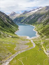 An alpine lake surrounded by green meadows and mountains, with a river and a hiking trail under a