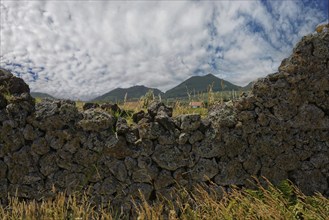 View of a lava stone wall with lush grasses, behind it green volcanic mountains under a cloudy sky,