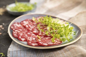 Slices of smoked cervelat salami sausage with spinach microgreen, salt and pepper on black concrete