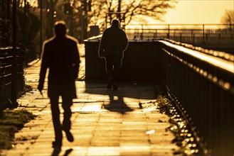 Man, elderly, walking on a pavement, a second man follows him, into the setting sun, long cast