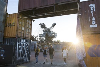 Festival visitors walk in the evening sun through a bridge made of containers in front of one of