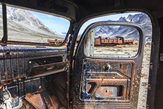 Interior view of a rusty car in front of steep mountains, remains of a US airbase from the Second