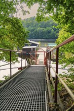 A staircase with metal railings leads down to a lake surrounded by green trees, Rappbodetalsperre,