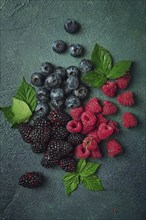 A mix of ripe berries, with leaves scattered on the table, raspberries, blackberries, blueberries,