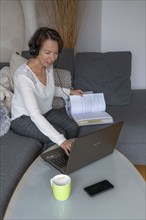 Woman, mid-50s, works from home, with laptop and communicates with colleagues via headset, home