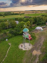 A tower in a rural area, surrounded by meadows and trees, shed near a dirt track under a cloudy