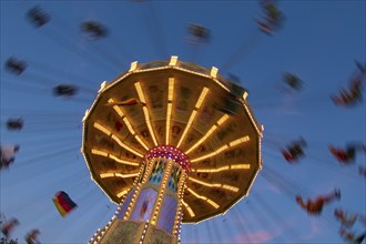 An illuminated carousel at dusk, spinning fast, in front of a blue sky with clouds and a Germany