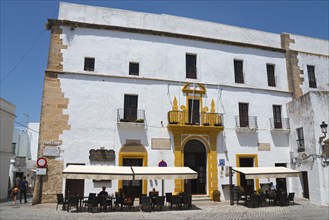 A historic building with a white façade and yellow entrance, including a street café with parasols,