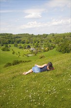 Woman lying on chalk grassland looking into deep green valley containing Rainscombe House, Oare