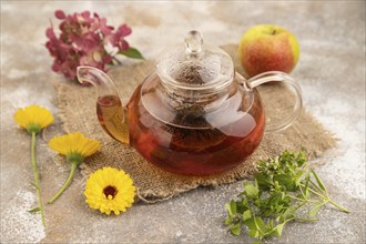 Red tea with herbs in glass teapot on brown concrete background and linen textile. Healthy drink