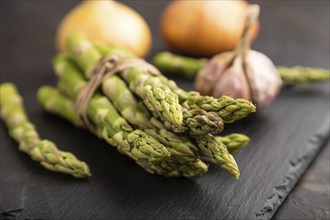 Bunch of fresh green asparagus, garlic, onion on slate board on black concrete background. Side