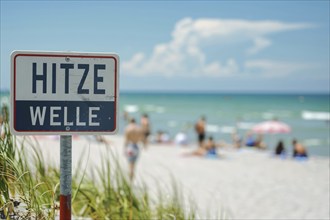 Warning sign with German text 'Hitzewelle' (Heat wave) in front of blurry sunny beach with people.