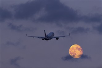 An aeroplane flies through the cloudy sky at night in front of a large, shining full moon, super