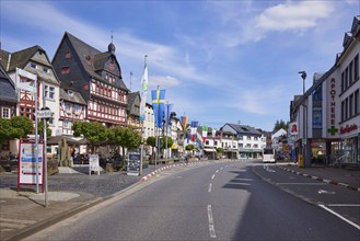 Main street with bus stop Adenau Markt, buildings, half-timbered houses, flags and trees under blue