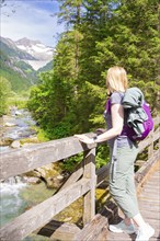 Woman with backpack looking at a river from a wooden bridge in a mountainous forest landscape,