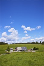 Farmstead with solar cells in a landscape with meadows and forest under a blue sky with cumulus