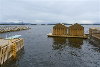 Outdoor sea pool by the fjord with small wooden sauna huts, overcast sky and calm water, Molde,