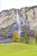 A waterfall flows down from high rocks, surrounded by trees in autumnal colours, Lauterbrunnen,