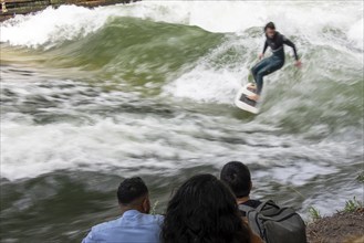 Surfer riding Eisbach wave, onlookers on the shore, Eisbach and wave in the English Garden are