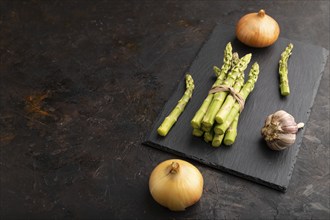 Bunch of fresh green asparagus, garlic, onion on slate board on black concrete background. Side