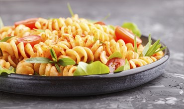 Fusilli pasta with tomato sauce, cherry tomatoes, lettuce and herbs on a black concrete background.