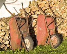 Two old red metal wheelbarrows leaning against pile wood logs in garden, Cherhill, Wiltshire,