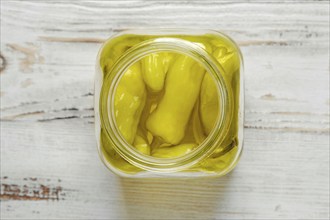 A glass jar of pickled peppers sits on a white wooden table. The jar is filled with whole, yellow