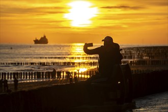 Sunset on the beach of Zoutelande, beach with wooden pile breakwaters, tourists, cargo ship sailing