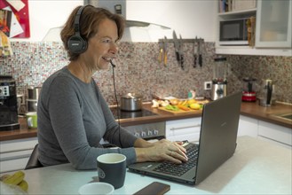 Woman, mid-50s, works from home, with laptop and communicates with colleagues via headset, home