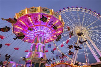 A lively funfair at dusk, illuminated by a large Ferris wheel and a colourful chain carousel,