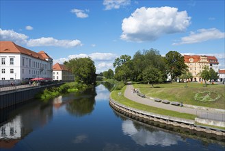 A quiet river with trees, park benches and historic buildings under a blue sky, Oranienburg, Havel