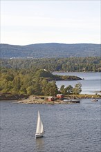 A single sailboat sails peacefully on the water, near a green island and wooded hills, Oslofjord,