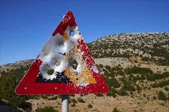 A damaged road sign with bullet holes in front of a mountainous landscape and blue sky, Lefka Ori,