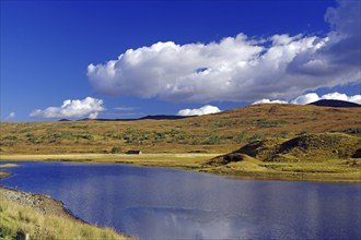 Lake surrounded by rolling hills, grass and a clear blue sky with white clouds Autumn, October,