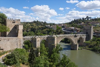 Old bridge over river in picturesque hilly landscape with fortress and blue sky with clouds, Puente