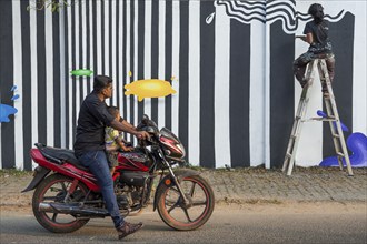 Father and son on motorbike watching Indian artist painting a wall during the International