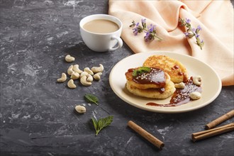 Cheese pancakes with caramel sauce on a beige ceramic plate and a cup of coffee on a black concrete