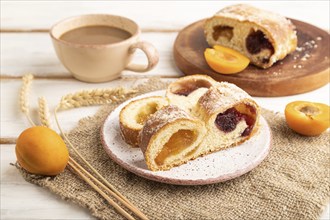 Homemade sweet bun with apricot jam and cup of coffee on white wooden background and linen textile.