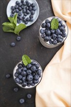 Yogurt with blueberry and chia in glass on black concrete background and orange linen textile. top