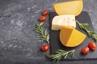 Various types of cheese with rosemary and tomatoes on black slate board on a black concrete