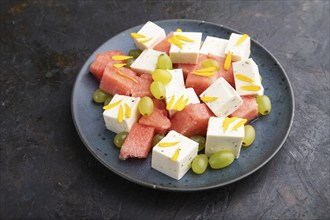 Vegetarian salad with watermelon, feta cheese, and grapes on blue ceramic plate on black concrete