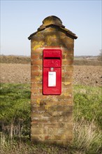 Scarlet red Post Office pillar box mounted in brick with fields in background, Hoo, Suffolk,