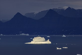 Evening light on an iceberg in a fjord in front of steep mountains, cloudy, autumn, Scoresby Sund,