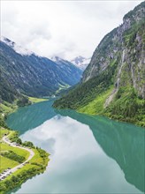 A clear, turquoise-coloured lake, surrounded by wooded mountain slopes and cliffs, under a cloudy