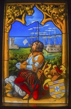 Stained glass window shows a knight in armour with a sailing ship and castles in the background,