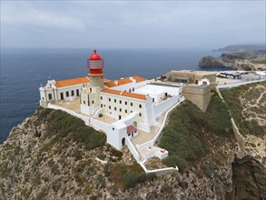 A red lighthouse on a rocky cliff, surrounded by white buildings, overlooking the ocean, aerial