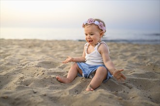 A smiling baby in summery clothes and flowers in her hair sits on the beach at sunset